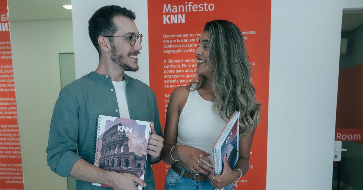Dois alunos KNN sorrindo com materiais didáticos em frente ao manifesto da escola