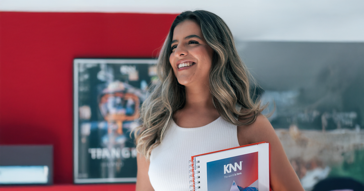 Aluna KNN sorrindo com caderno da escola em ambiente interno colorido e com o livro da KNN Idiomas.