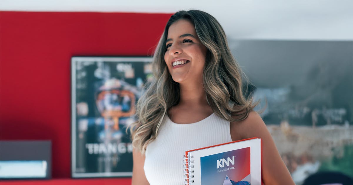 Aluna KNN sorrindo com caderno da escola em ambiente interno colorido