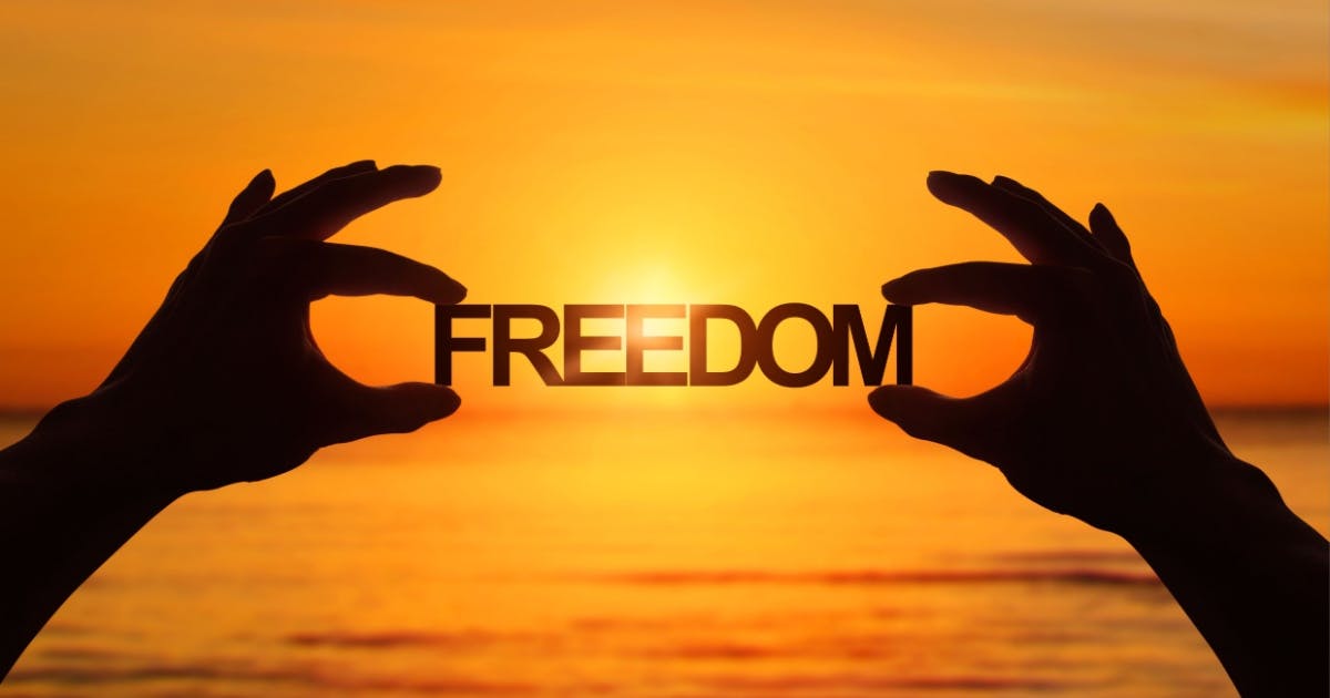Imagem de duas mãos segurando a palavra "FREEDOM". Ao fundo, uma paisagem com o mar e pôr do sol.