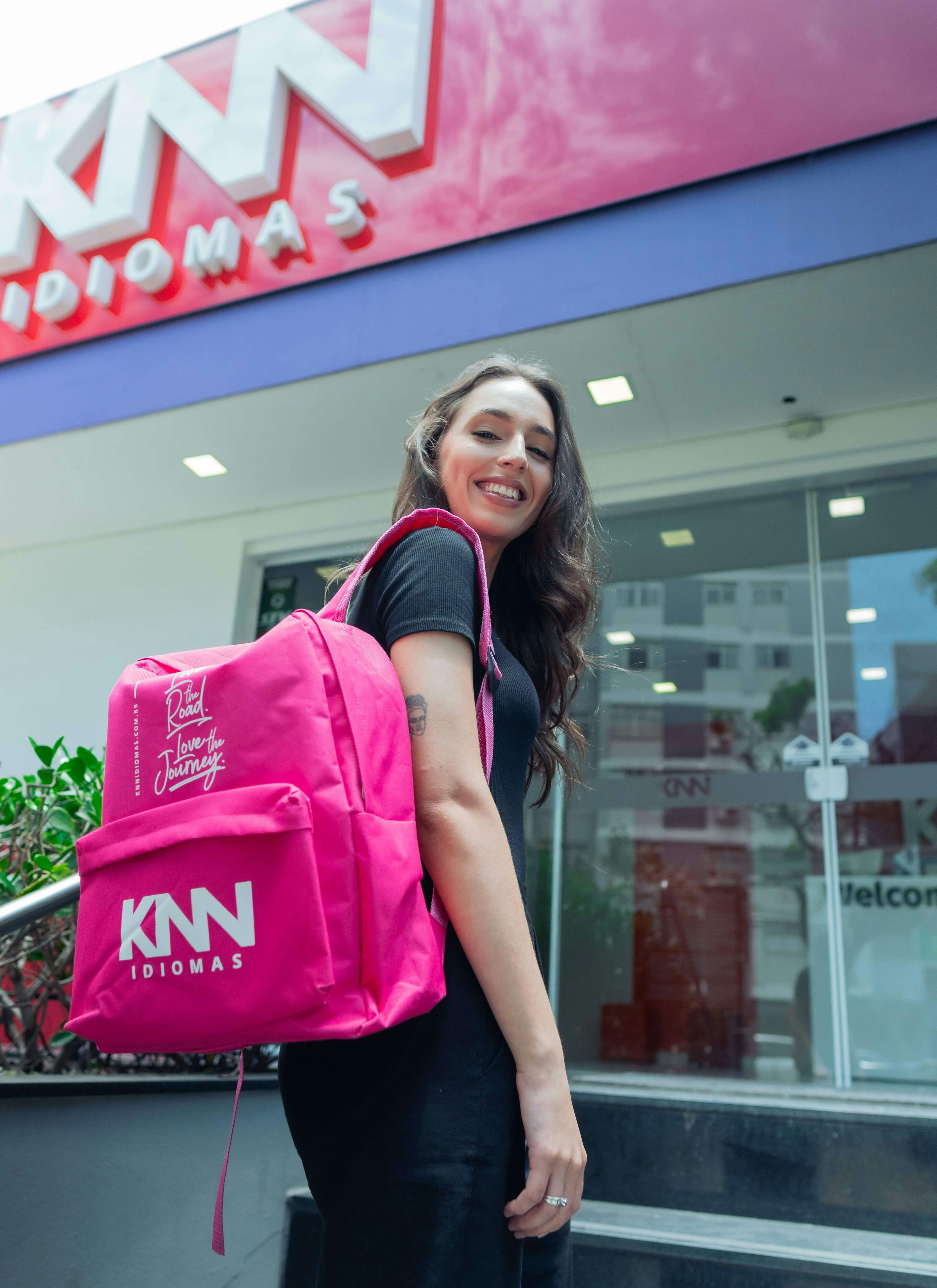 Mulher de mochila rosa e sorrindo, em frente a escola KNN Idiomas.