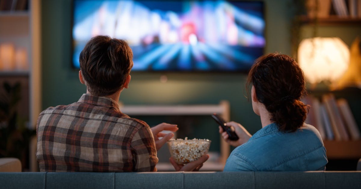 Um homem e uma mulher de costas assistindo um filme na TV. A mulher está com o controle na mão.