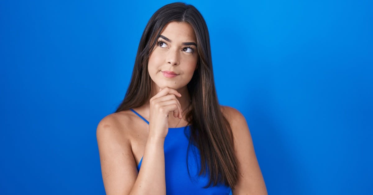 Foto de uma mulher com regata azul, mão no queixo e expressão de dúvida.