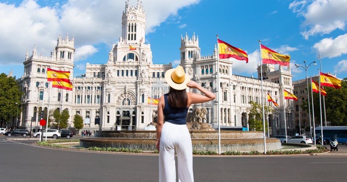 Mulher de costas, e com chapéu virada de costas e olhando para um castelo com várias bandeiras da Espanha na frente.