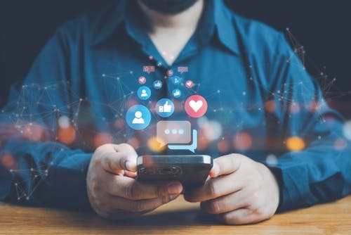 Pessoa com o celular na mão e hologramas de "likes" e "corações" saindo do celular em formato de holograma.