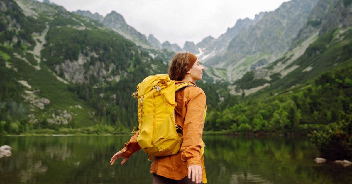 Mulher com os braços abertos e um mochila nas costas. Ao fundo, uma paisagem de natureza.