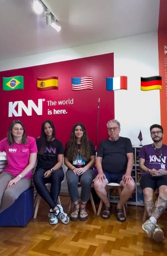 Foto dos professores da KNN Idiomas sentados em cadeiras e as bandeiras de Brasil, Espanhol, EUA, França e Alemanha em cima de suas cabeças.
