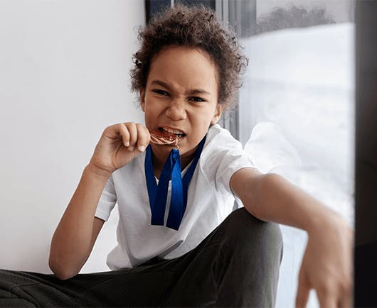 Uma criança de camiseta branca comemorando com uma medalha. Ela está mordendo a medalha.