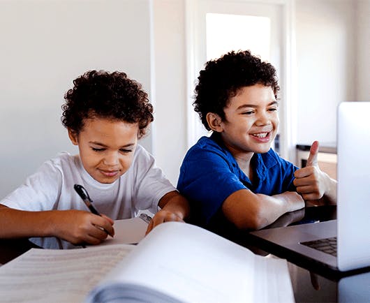 Duas crianças sentadas, uma escrevendo em um livro e a outra olhando para um notebook.