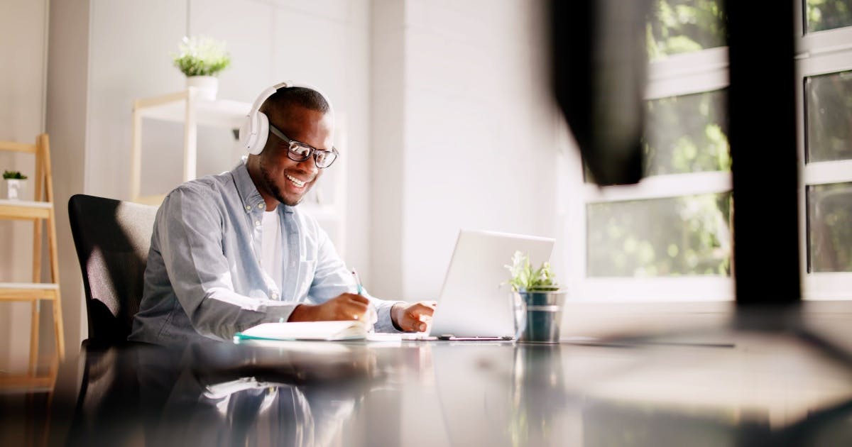Pessoa com fone de ouvido, sentada em uma mesa, sorrindo e com um notebook em cima da mesa.