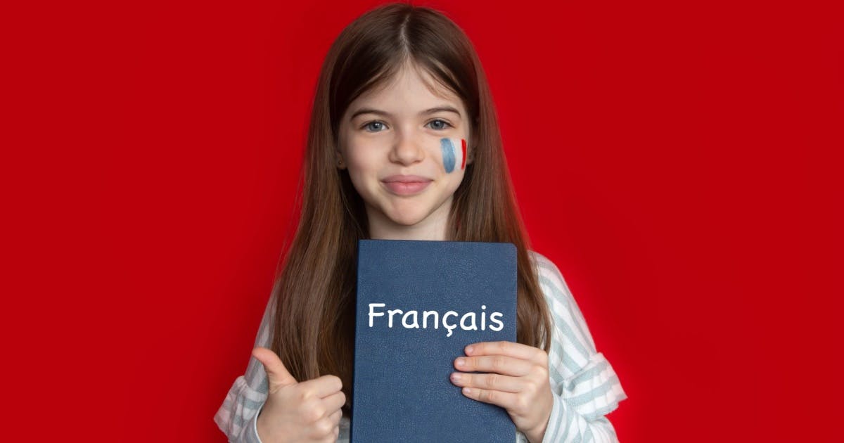 Criança com um livro de francês na mão esquerda e, com a direita, fazendo um sinal de positivo. No seu rosto, está desenhado uma bandeira da frança.