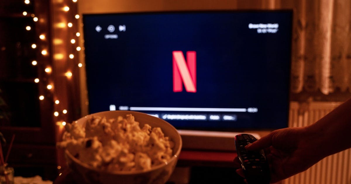 Foto de uma pessoa com um balde de pipoca e um controle na mão apontando para uma TV com a tela da Netflix.