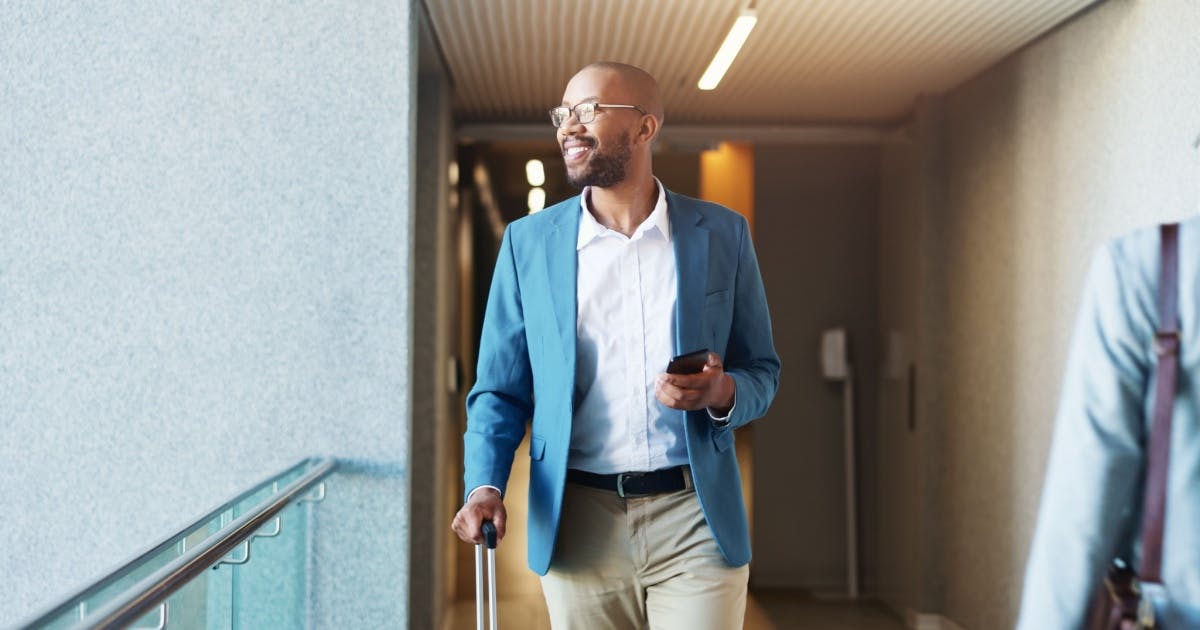 Homem com roupa social e uma mala de rodinhas na mão direito, na esquerda, está com um celular.