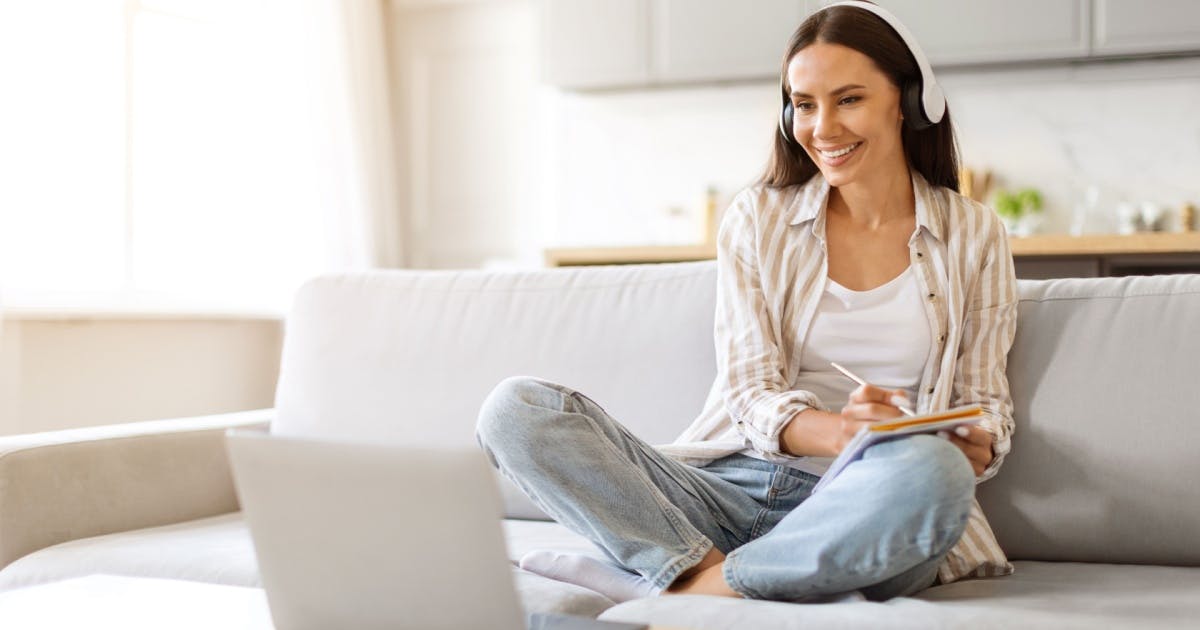 Mulher sentada no sofá, com fones de ouvido e um notebook em uma mesinha.