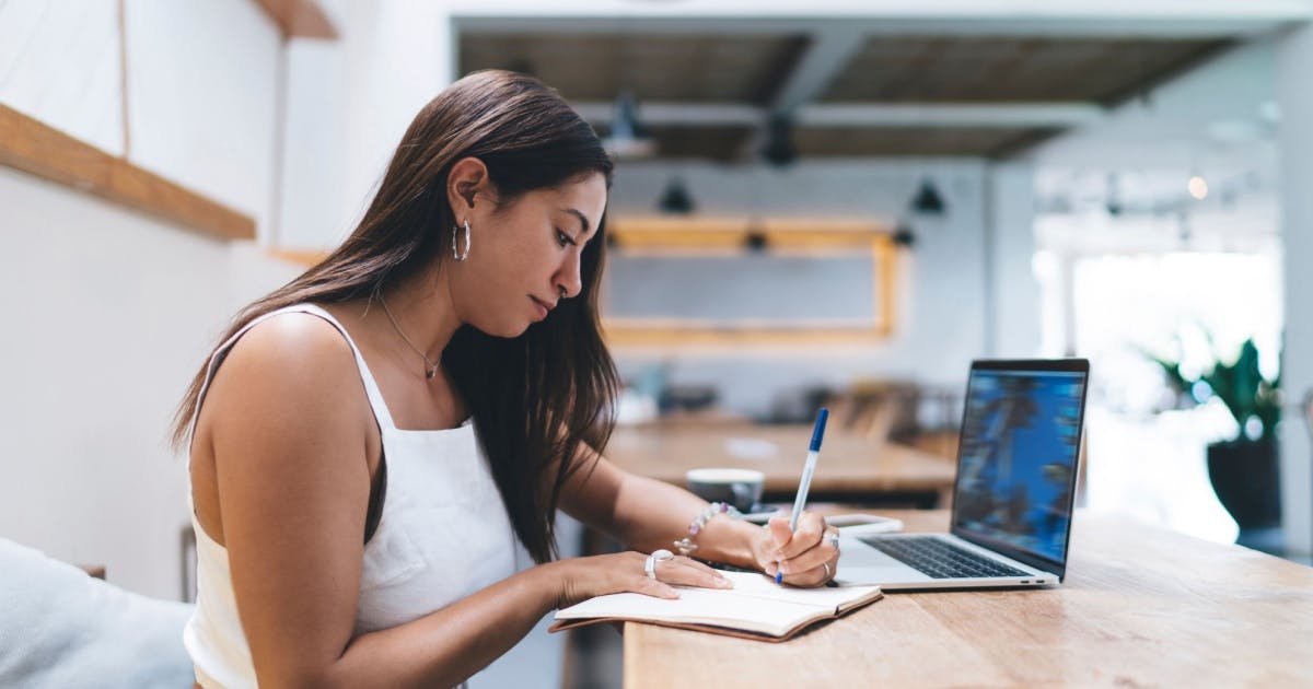 Mulher sentada, escrevendo e com um notebook aberto.
