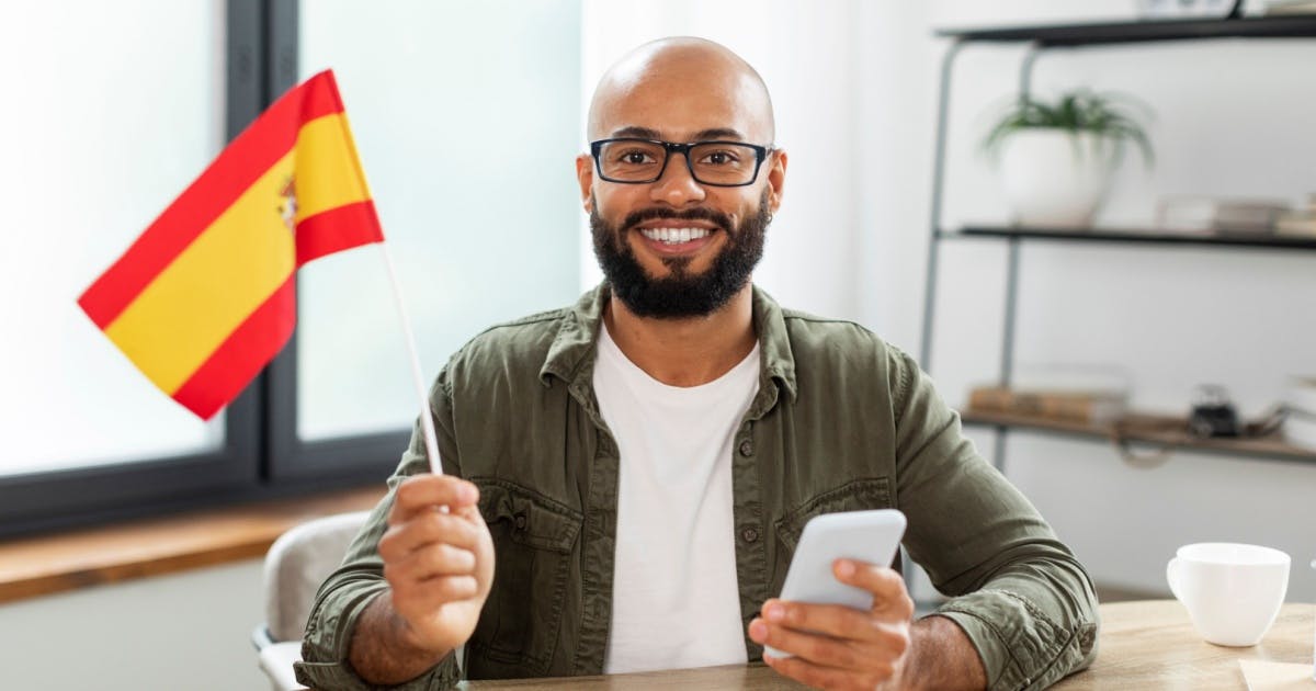 Homem de óculos sorrindo, com um celular na mão esquerda, e a bandiera na mão direita. 