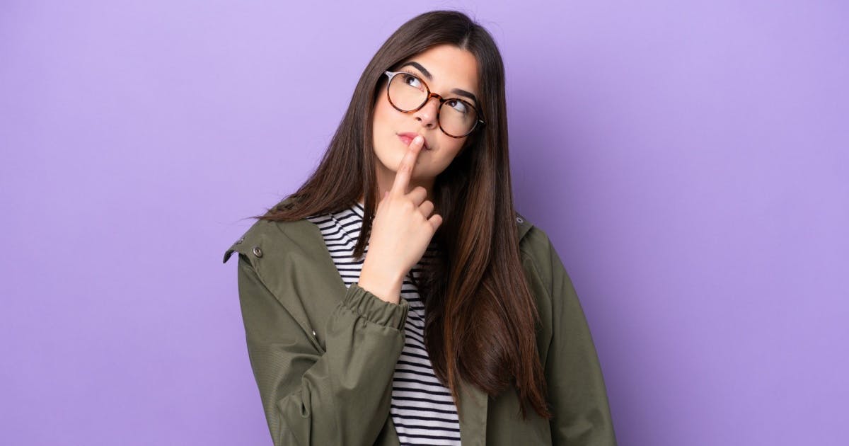 Mulher de cabelo preto, comprido e liso, usando um óculos, com o dedo indicador na boca, olhando para cima e com expressão de dúvida.