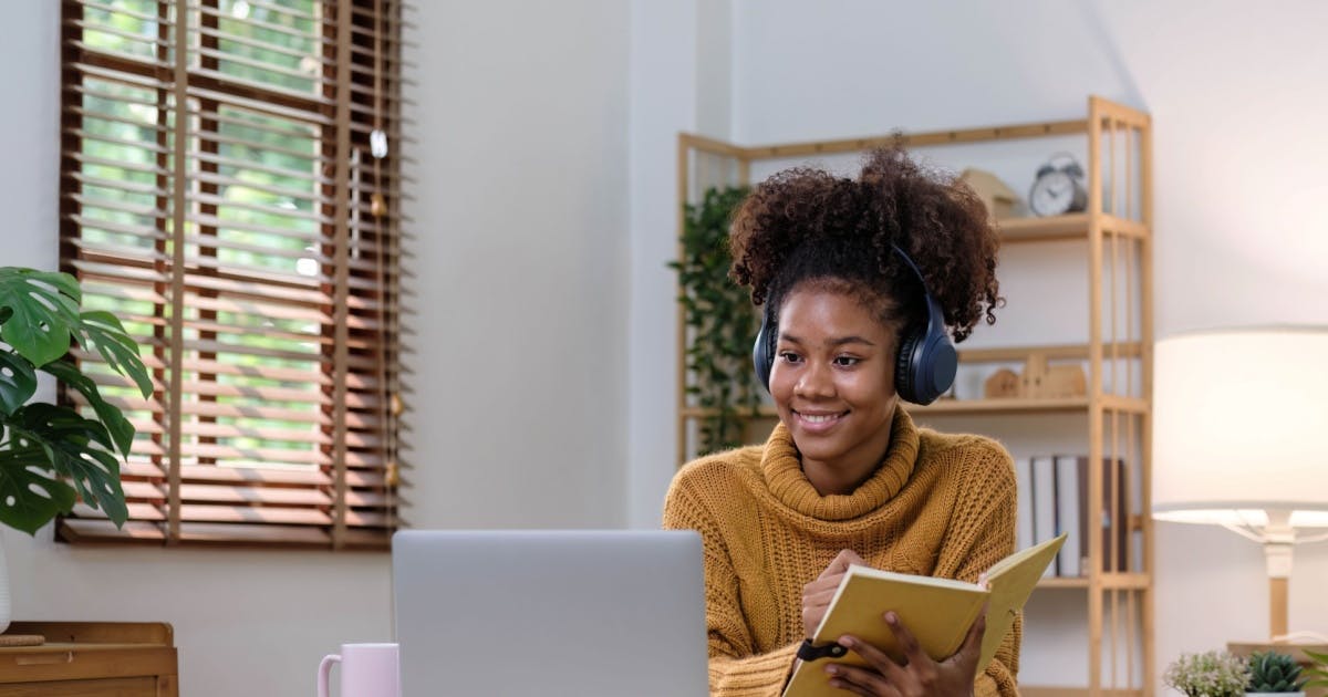 Mulher com um livro na mão, fone de ouvido e olhando para um notebook. Ela está sorrindo.
