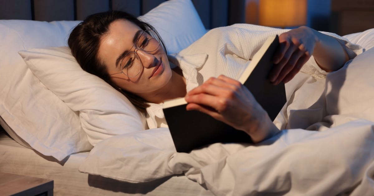 Mulher com óculos, deitada na cama e lendo.