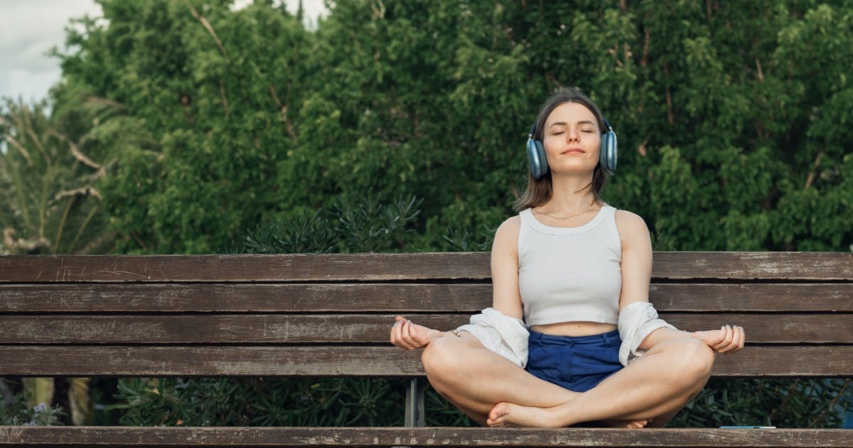 Mulher com fones de ouvido sentada em um banco de praça e meditando.
