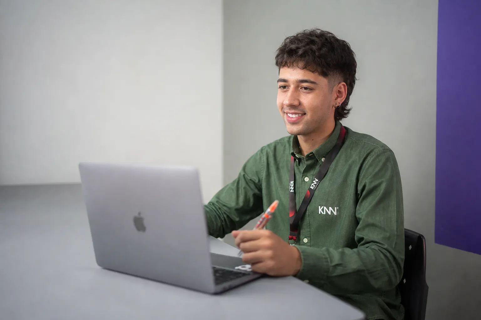 Homem sentado olhando para a tela de um notebook e uma caneta na mão esquerda.