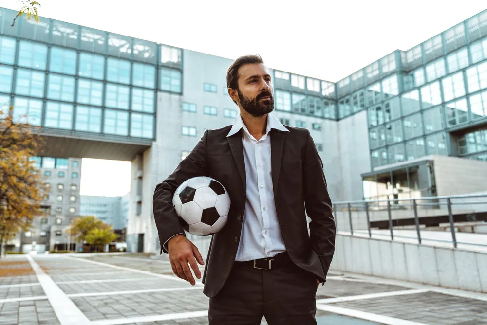 Homem de terno segurando uma bola de futebol olhando para o horizonte.