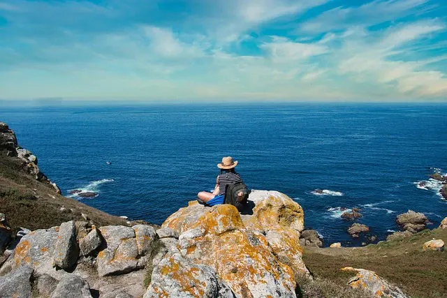 Pessoa sentada sobre rochas à beira do mar, usando chapéu de palha, com o oceano ao fundo e céu azul com nuvens leves, cenário costeiro natural