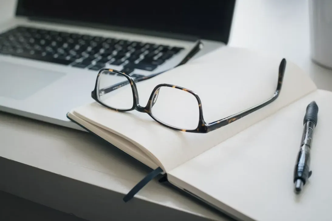 Óculos de armação tartaruga sobre um caderno aberto com caneta ao lado, em uma mesa de escritório com notebook e teclado desfocados ao fundo. Foto em ambiente corporativo.
