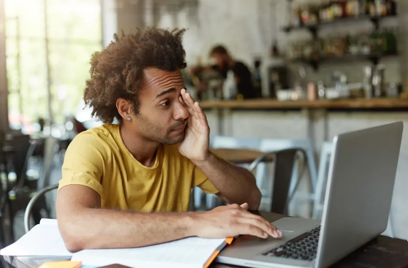 Homem jovem com cabelo afro encaracolado em uma cafeteria, com expressão pensativa e mão na orelha, usando laptop e consultando documentos sobre a mesa.