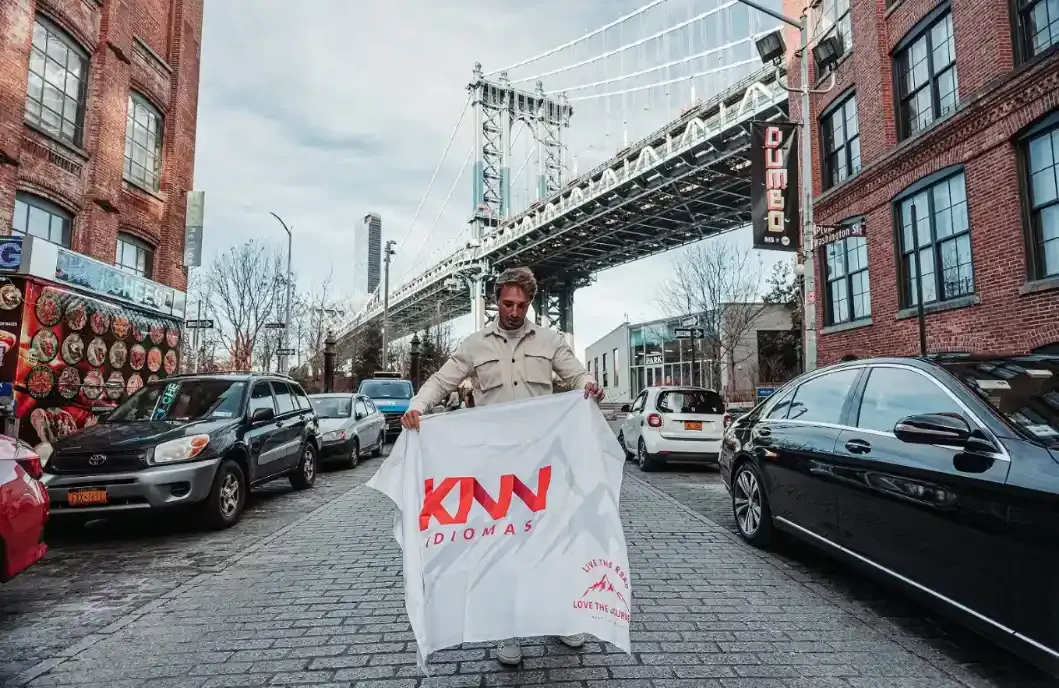 Homem segurando uma sacola branca com o logotipo “KNN Idiomas” em frente a um viaduto/ponte antiga entre prédios de tijolos, ao lado de carros estacionados na rua.
