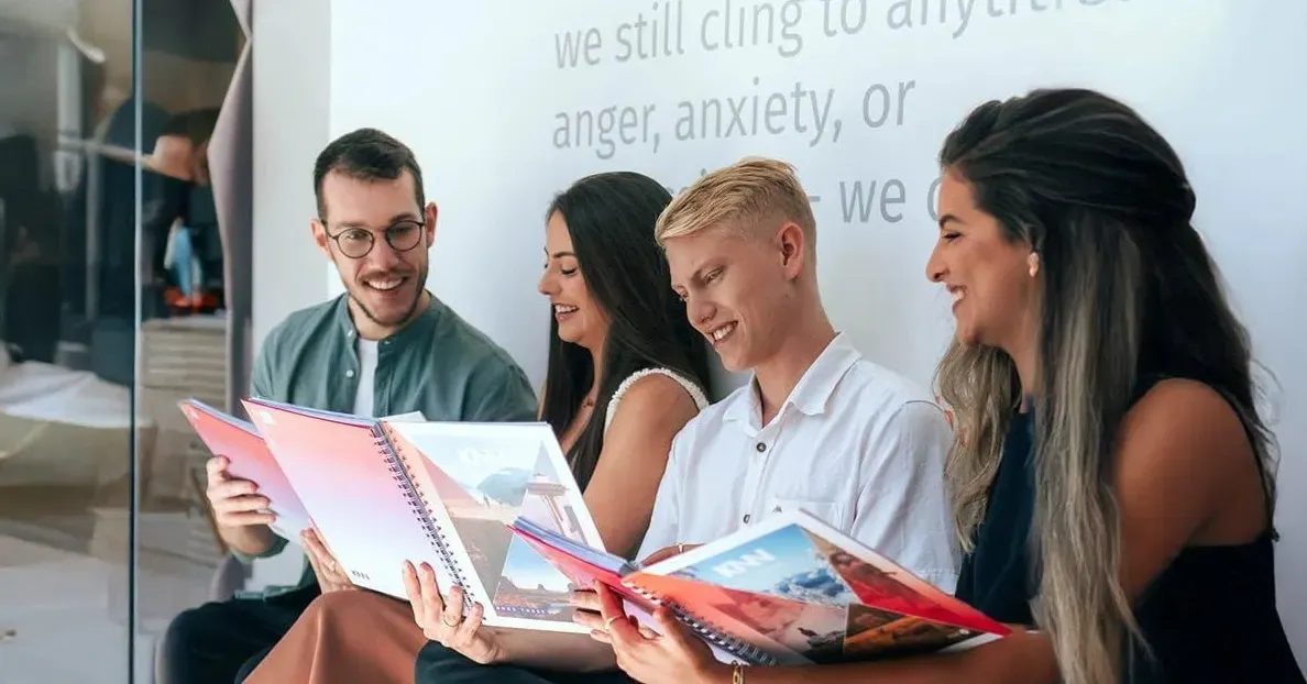 Equipe de trabalho em escritório, sorrindo e folheando revistas ou folders abertos, com um quadro ao fundo e texto sobre emoções como raiva e ansiedade.