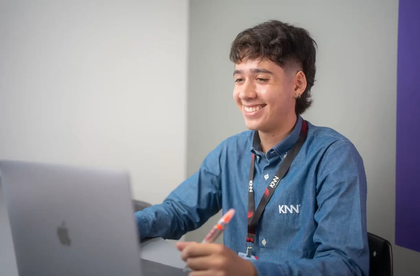 Jovem sorrindo e trabalhando em um computador em um ambiente de escritório, usando camisa jeans e crachá, com expressão de atenção durante uma conversa ou atendimento.