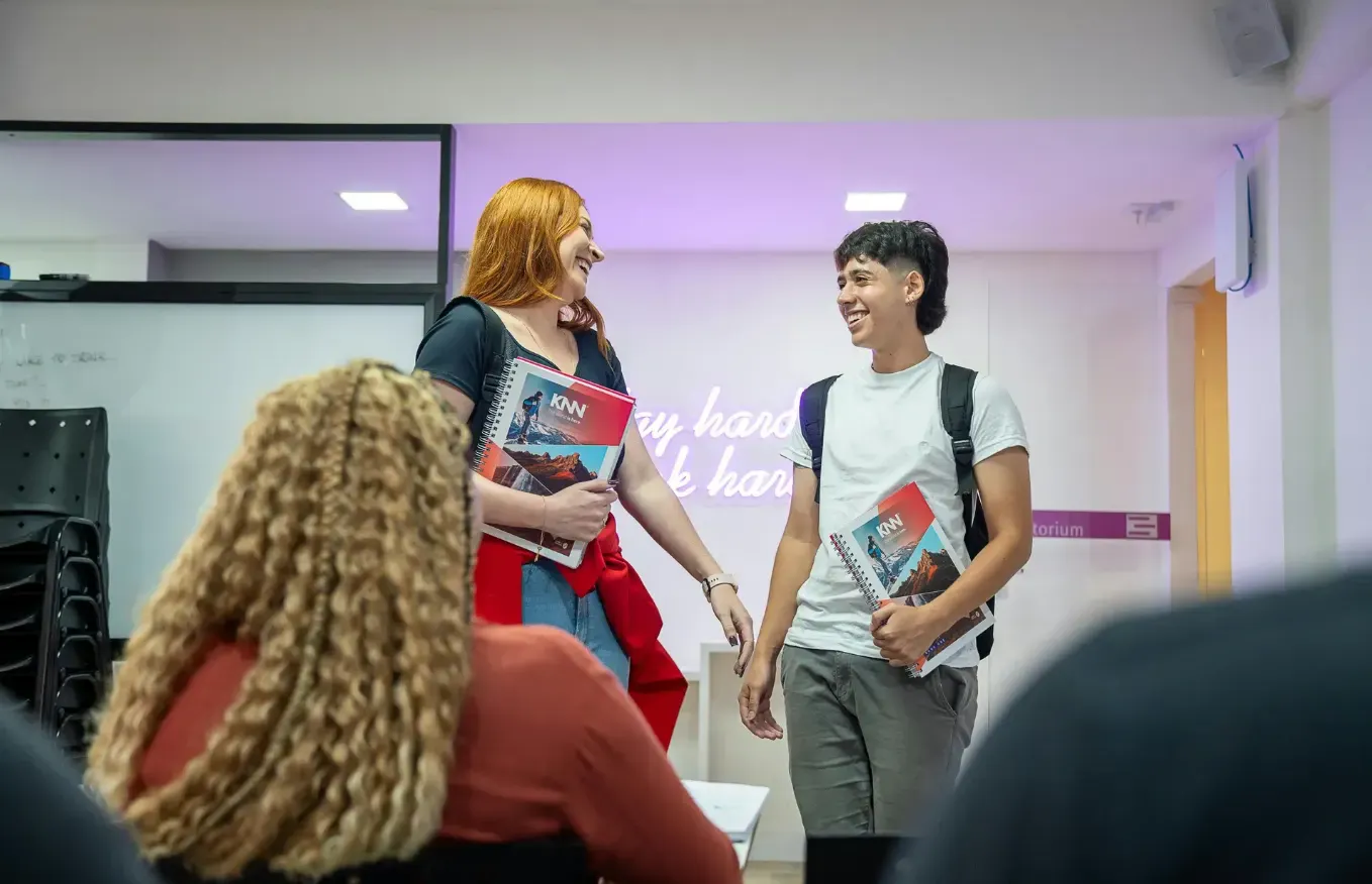 Alunos em sala de aula conversam e caminham sorrindo, segurando apostilas e mochila, em um ambiente educacional com quadro branco ao fundo e luzes no teto.