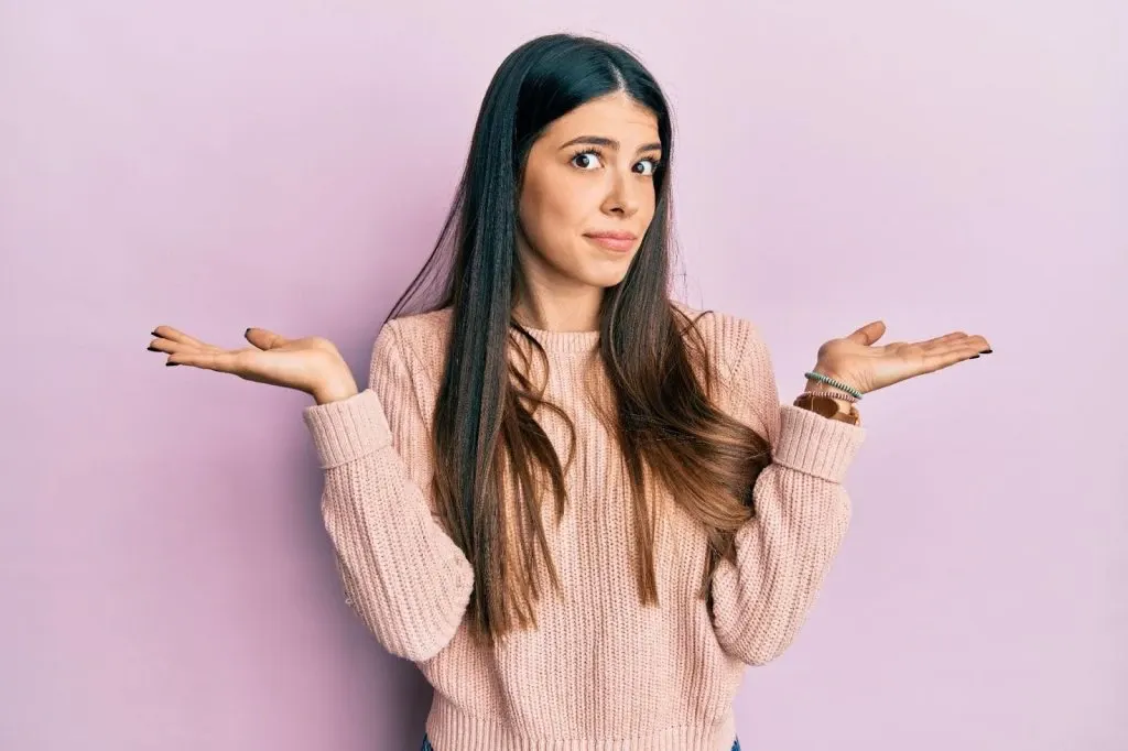 Mulher jovem com cabelo longo e vestido de tricô bege em fundo rosa, com as mãos abertas e expressão confusa, estilo casual em estúdio.