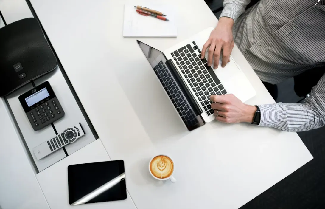 Pessoas em ambiente de escritório usando um notebook para trabalhar com as mãos no teclado, com calculadora, tablet e café sobre a mesa, e documentos ao fundo.