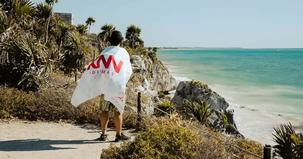 Homem com camisa branca da marca DIMASA e short camuflado caminha na beira de uma praia de águas turquesa, com coqueiros e vegetação nas falésias, sob céu azul.