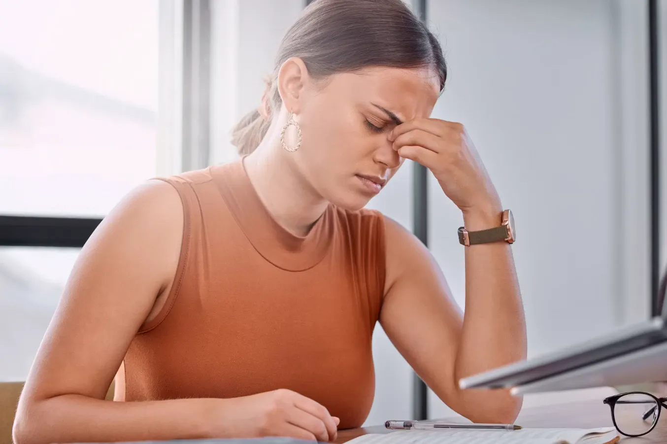 Mulher sentada em ambiente corporativo, com expressão cansada e mão no rosto, enquanto olha para documentos e celular sobre a mesa, sugerindo estresse no trabalho.