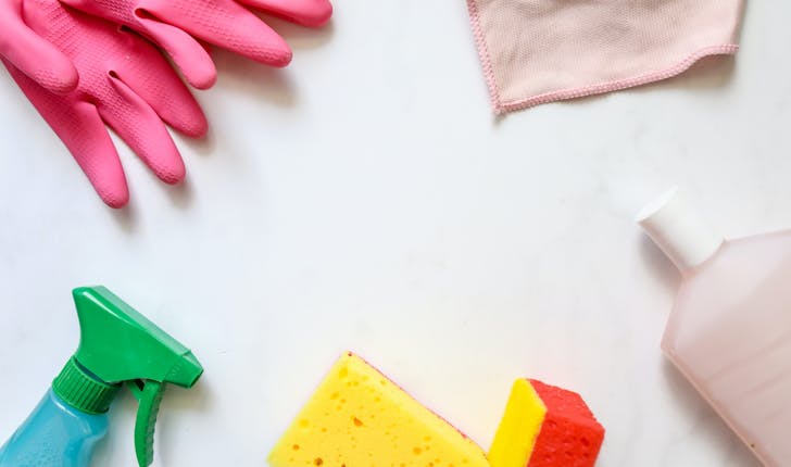 cleaning utensils on a table: pink cleaning gloves, a blue and green bottle with cleaning product, yellow sponges and and a pink cloth