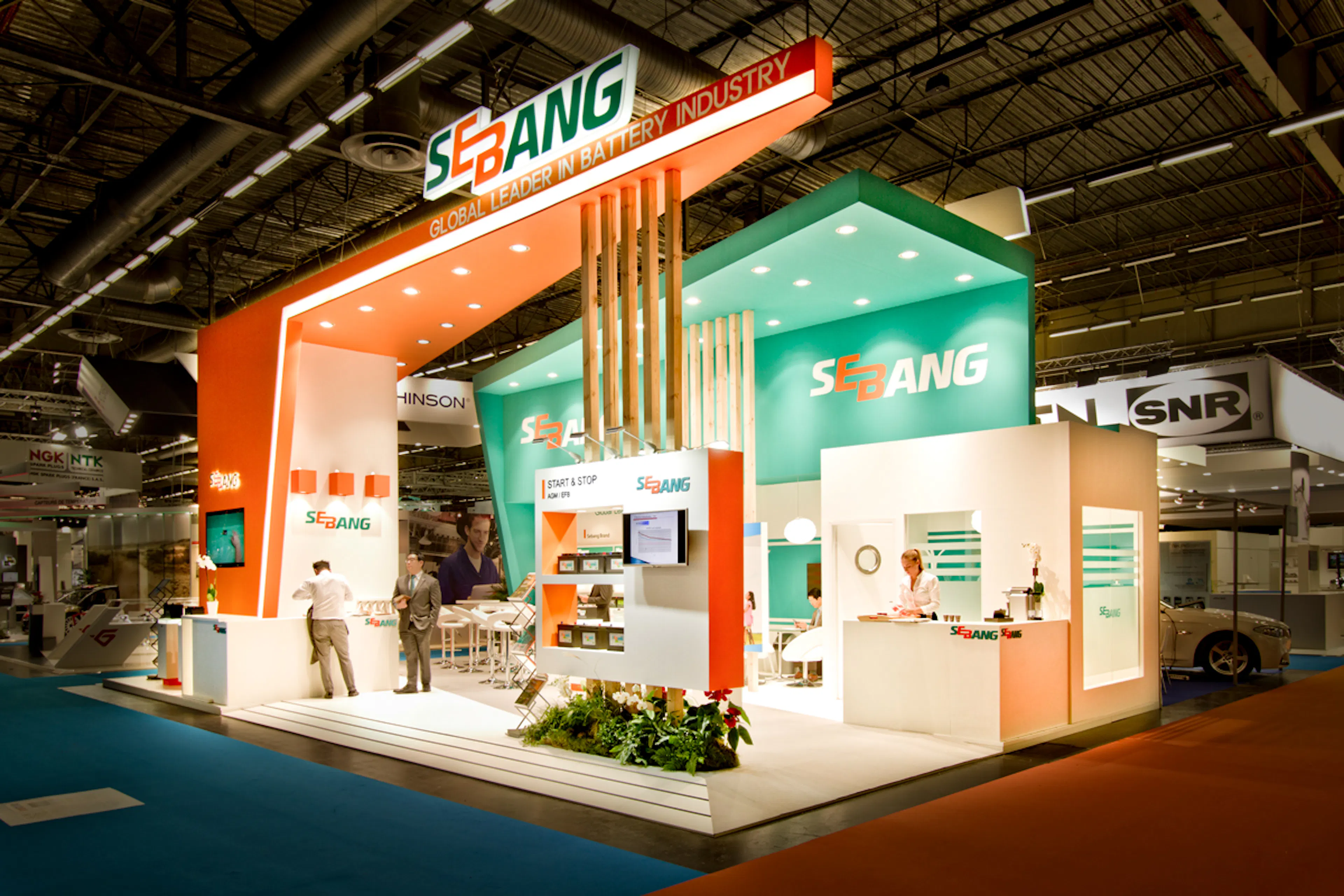 Exhibition stand for Sebang, a battery manufacturer, at a trade fair, featuring an orange and teal color scheme with the tagline "Global Leader in Battery Industry," an internal meeting room, product display shelving, and a reception counter with a staff member.