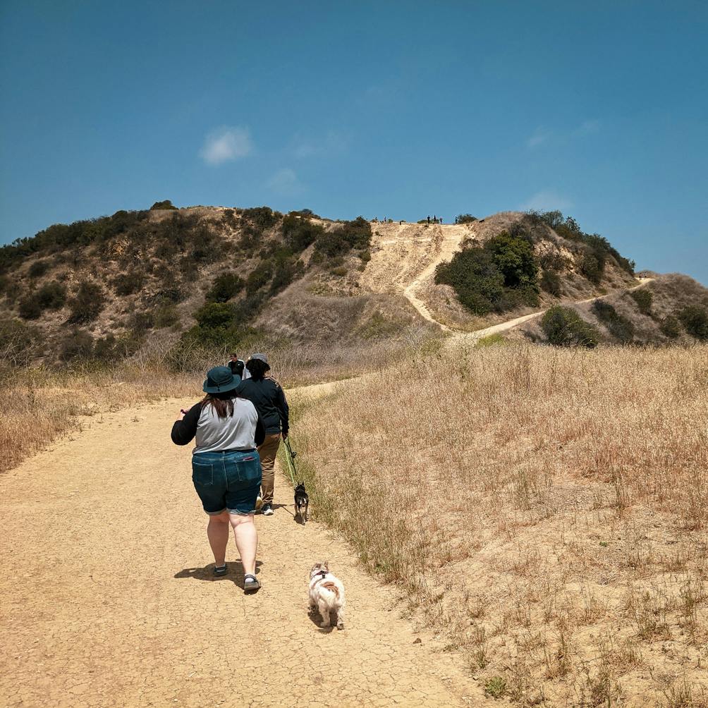 Two hikers with a dog going along a wide dirt trail at Upper and Lower Canyonback Trails in Los Angeles County 