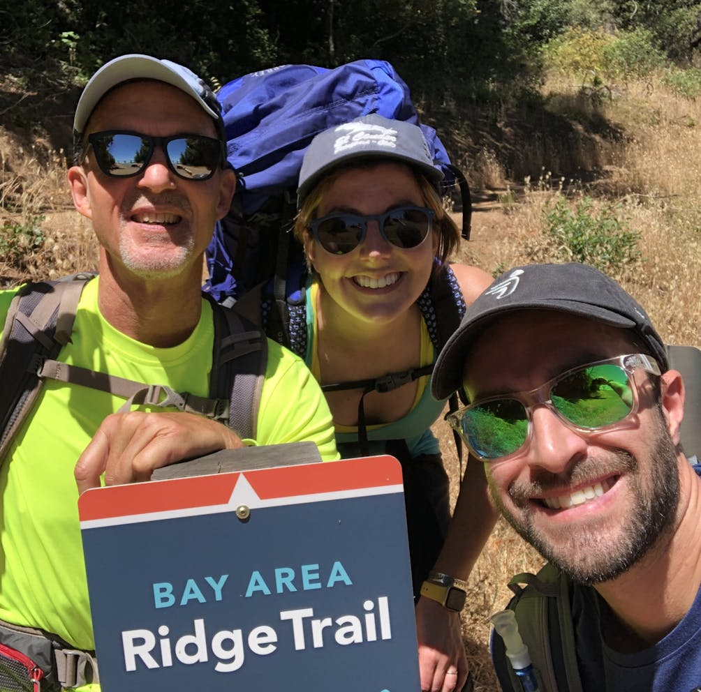 Three hikers smiling at the camera in front of the Bay Area Ridge Trail 