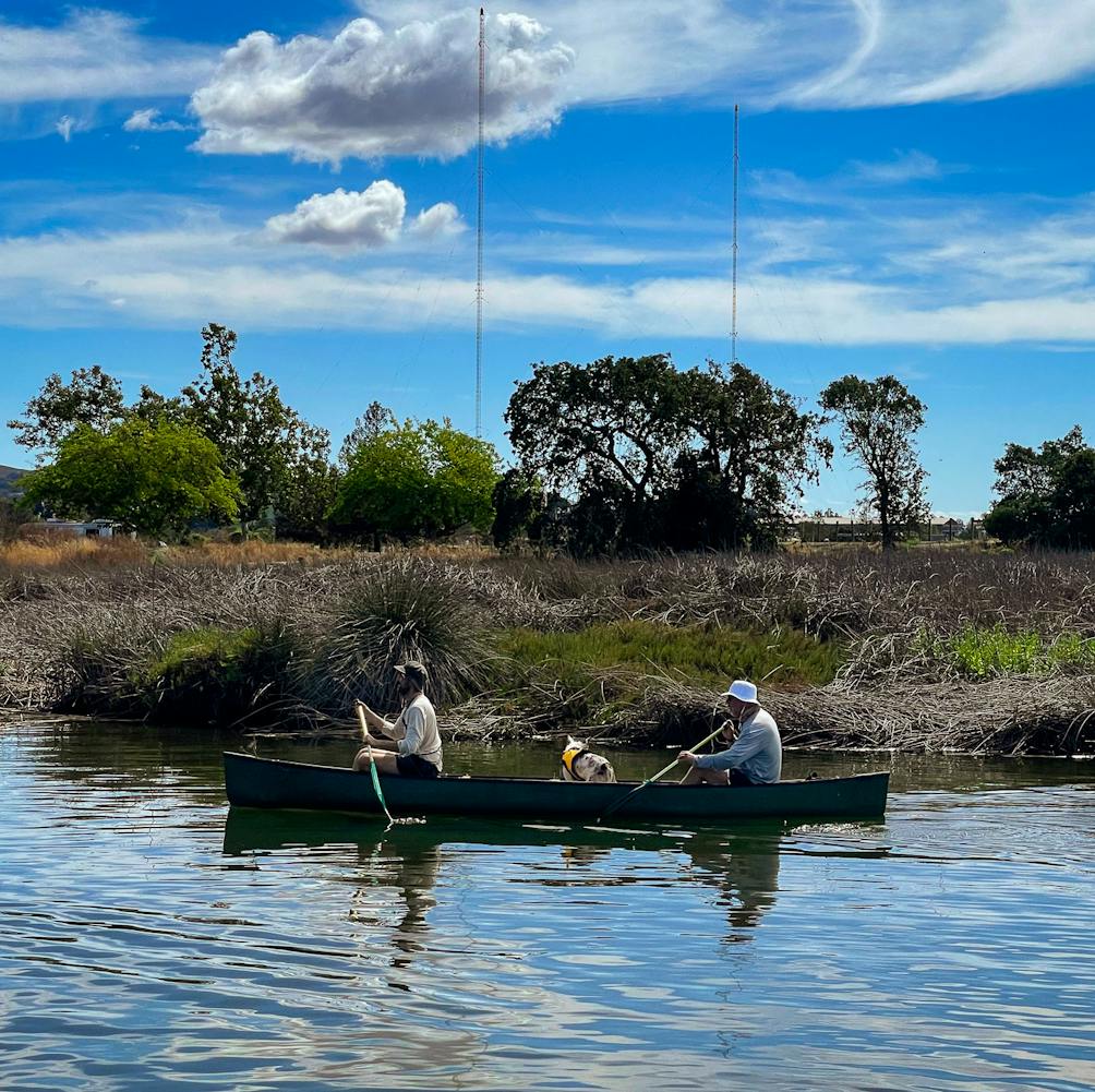 Two people and a dog in a canoe paddling along the Napa River 