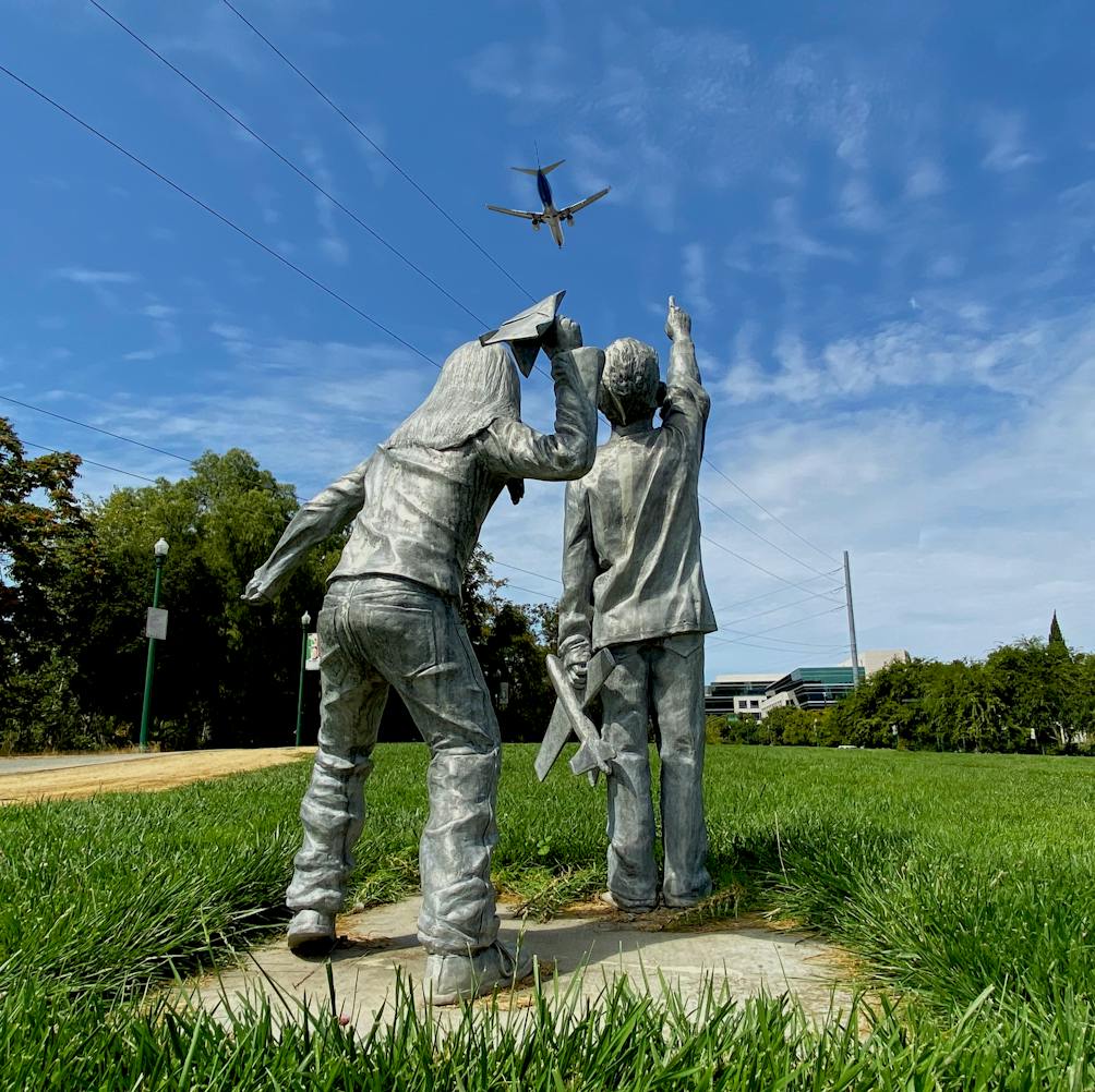 Sculpture of two kids pointing at the sky near the San Jose Airport with planes flying overhead