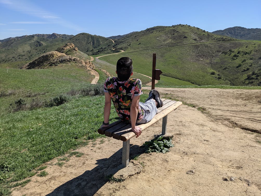Hiker taking a break sitting on a bench overlooking rolling green hills and a winding hiking trail at Long Canyon in Simi Valley 