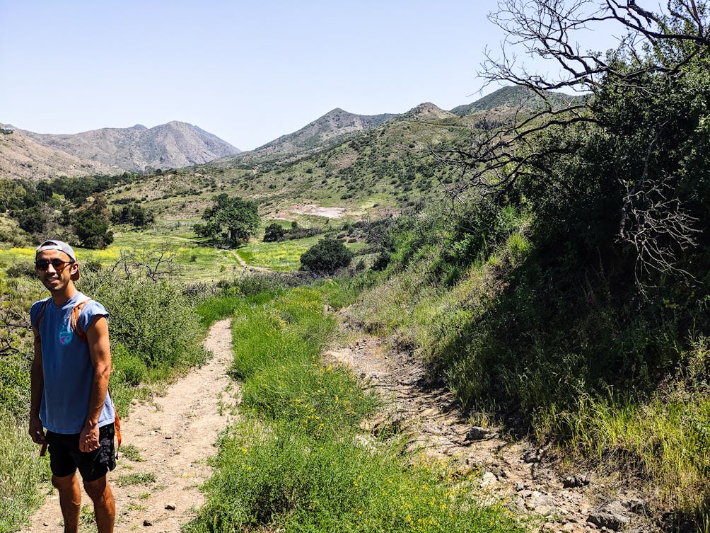 A hiker smiling at the camera on a trail with mountains in the background at Triunfo Creek Park in Southern California