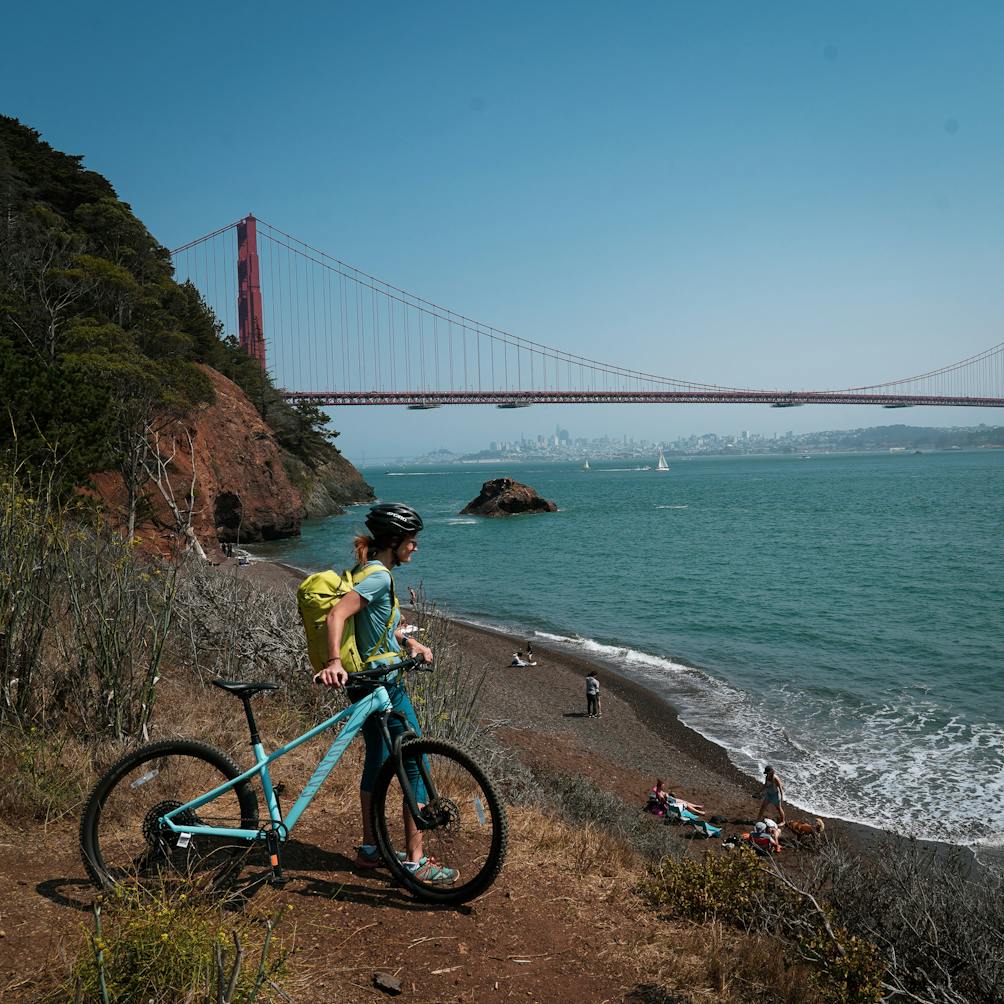 A biker with a yellow backpack standing at a vantage just above Kirby Cove Beach with the Golden Gate Bridge in the background 