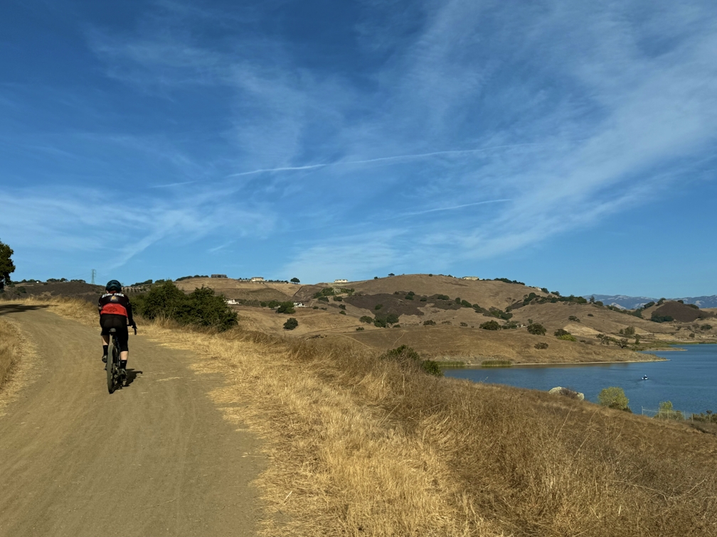 Person riding bike on wide dirt trail next to a lake at Calero County Park