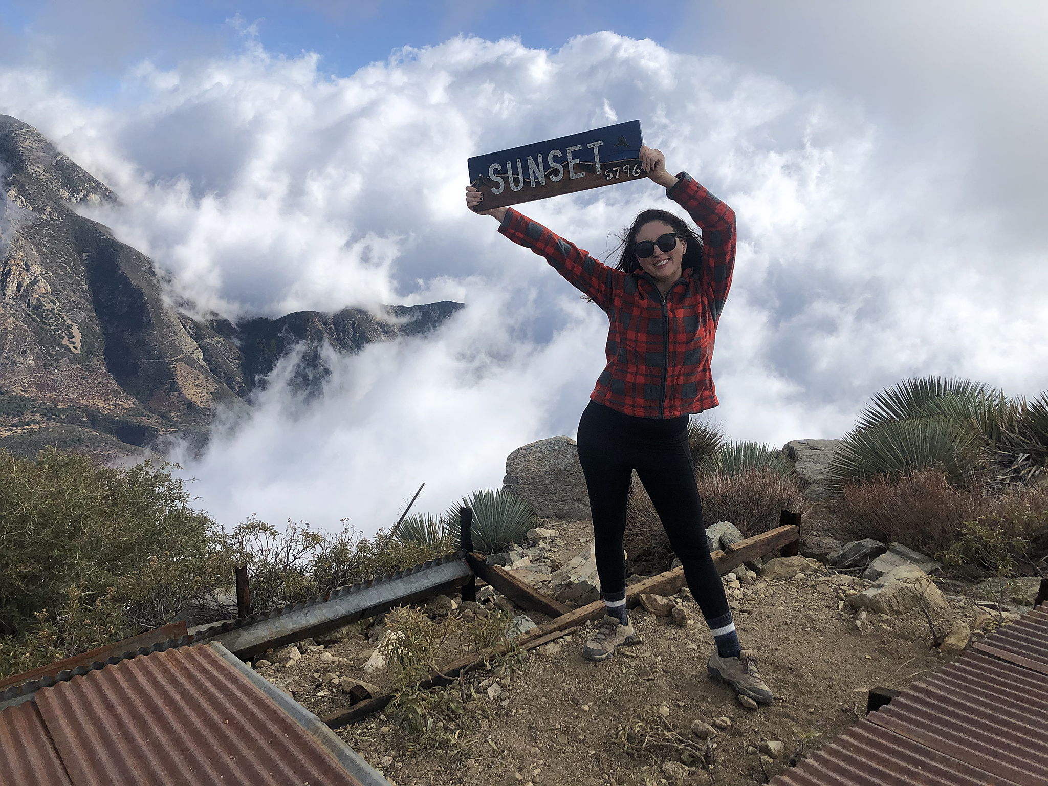 Woman holding a wood sign saying Sunset at Sunset Peak in the San Gabriel Mountains 