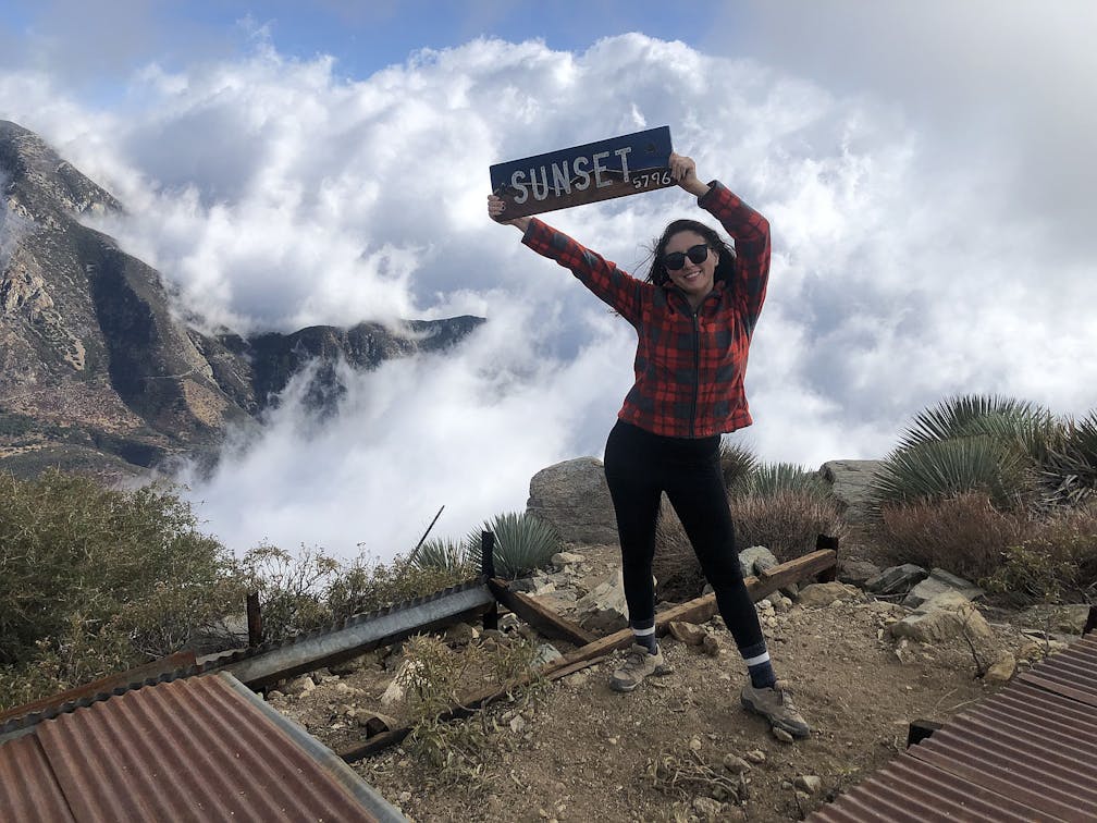 Woman holding a wood sign saying Sunset at Sunset Peak in the San Gabriel Mountains 