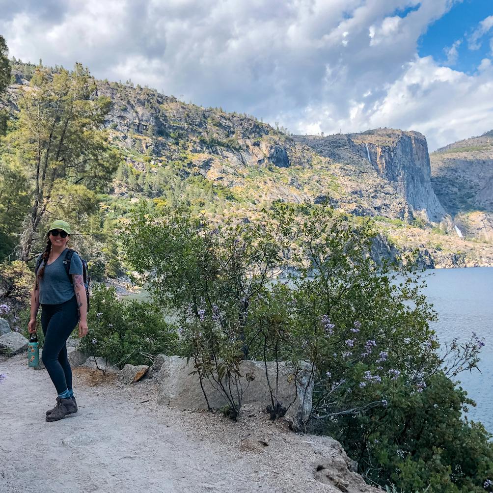 Woman smiling on the hiking trail with a lake in the background at Hetch Hetchy in Yosemite